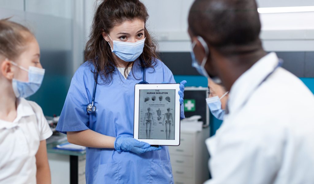 Pediatrician nurse with protective face mask against covid19 holding tablet computer with bones radiography on screen. African american doctor explaining medical expertise in hospital office